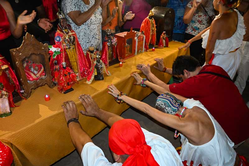 Bajo los árboles , las personas de todas las edades, vestidas de rojo y blanco, esperan ansiosas el tradicional baile y los toques de tambor que saludan al Santo Kambá. En este rincón del parque, lejos del bullicio del recital, se concentra la verdadera devoción, donde el culto se celebra con una intensidad única.